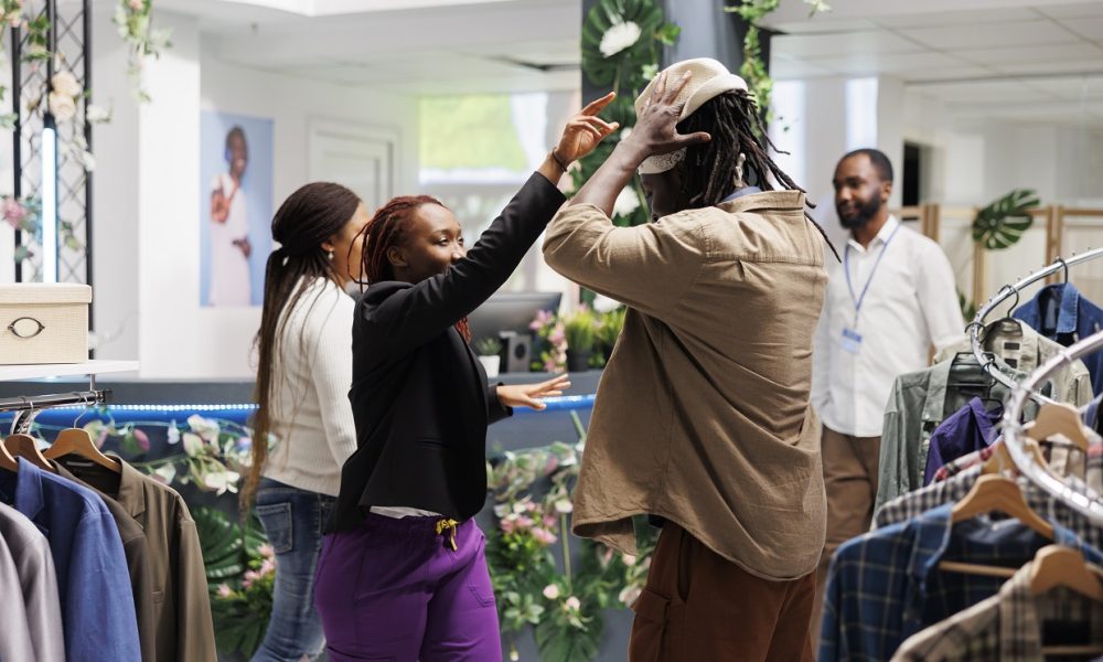 Stylish african american friends trying on hat and having fun together in clothing store. Man and woman customers checking out accessories and choosing outfit in shopping mall